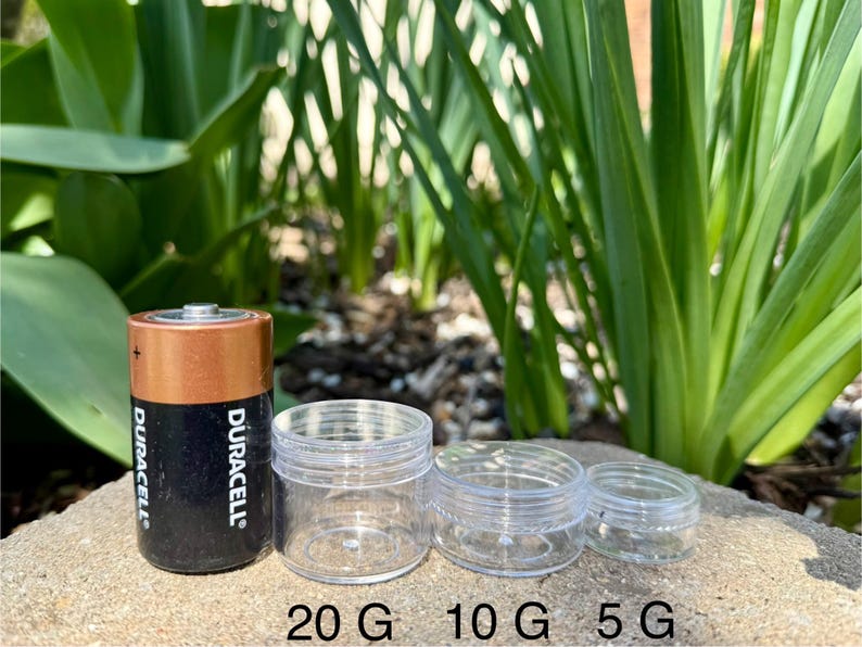 Op de afbeelding: Drie transparante plastic containers met deksels, gelabeld 20 G, 10 G en 5 G. Een Duracell batterij staat naast de containers voor groottevergelijking.