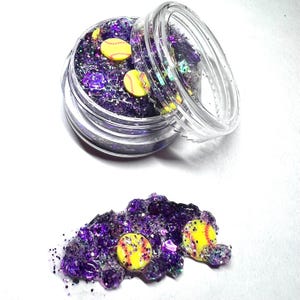 May include: A small clear jar filled with purple glitter and yellow softball charms. The jar is open and some of the glitter and charms have spilled out onto a white surface.