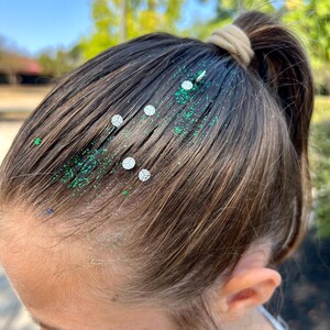 May include: A person with brown hair has green glitter and small white volleyball stickers in their hair. The hair is pulled back in a ponytail.