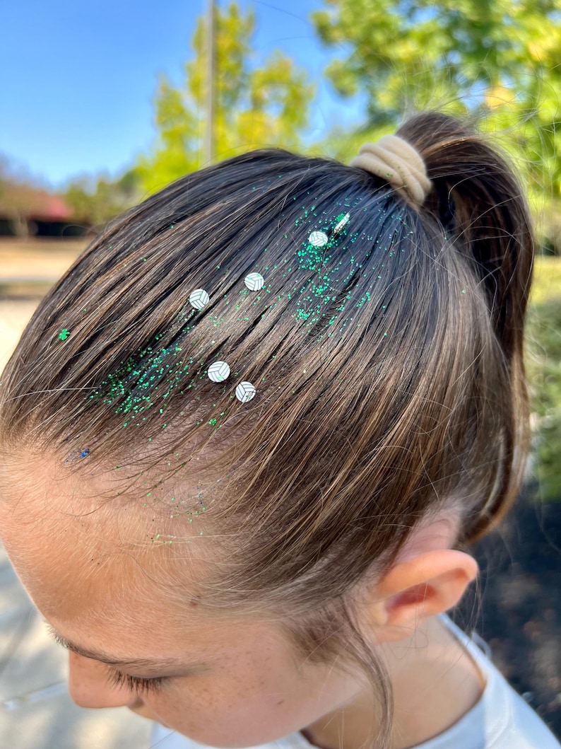 May include: A person with brown hair has green glitter and small white volleyball stickers in their hair. The hair is pulled back in a ponytail.