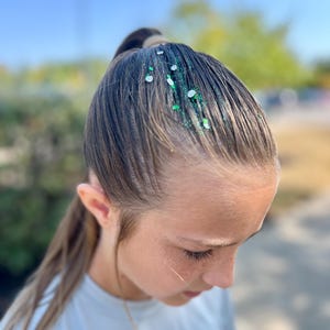 May include: A young person with long brown hair pulled back in a ponytail. Green and silver glitter is sprinkled on the hair.