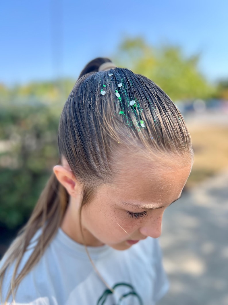 May include: A young person with long brown hair pulled back in a ponytail. Green and silver glitter is sprinkled on the hair.