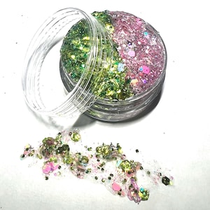 Op de afbeelding: Een klein, doorzichtig potje gevuld met groene en roze glitter. Het potje is open en wat glitter is op een wit oppervlak gevallen.