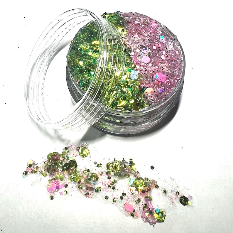Op de afbeelding: Een klein, doorzichtig potje gevuld met groene en roze glitter. Het potje is open en wat glitter is op een wit oppervlak gevallen.