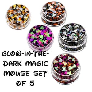 May include: A set of five clear plastic jars filled with colorful craft glitter in various shapes, including small mouse head silhouettes. The text GLOW-IN-THE-DARK MAGIC MOUSE SET OF 5 is printed in black on the left side of the image.