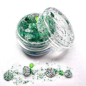 May include: A clear jar filled with green glitter and white volleyball charms. The jar is open and some of the glitter and charms have spilled out onto a white surface.