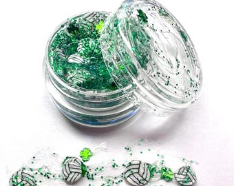Puede incluir: Un frasco transparente lleno de purpurina verde y abalorios de voleibol blancos. El frasco está abierto y parte de la purpurina y los abalorios se han derramado sobre una superficie blanca.