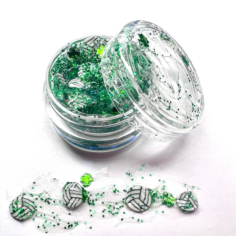May include: A clear jar filled with green glitter and white volleyball charms. The jar is open and some of the glitter and charms have spilled out onto a white surface.