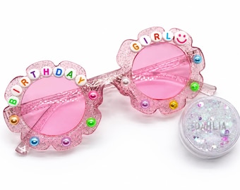 May include: Pink glittery flower-shaped sunglasses with pink lenses. The frames are decorated with colorful beads spelling BIRTHDAY GIRL and a smiley face. A small round container of clear iridescent glitter labeled DAHLIA is included.