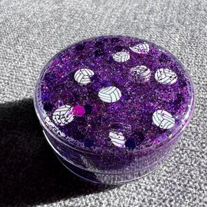 May include: A clear round container filled with purple glitter and white volleyball designs.