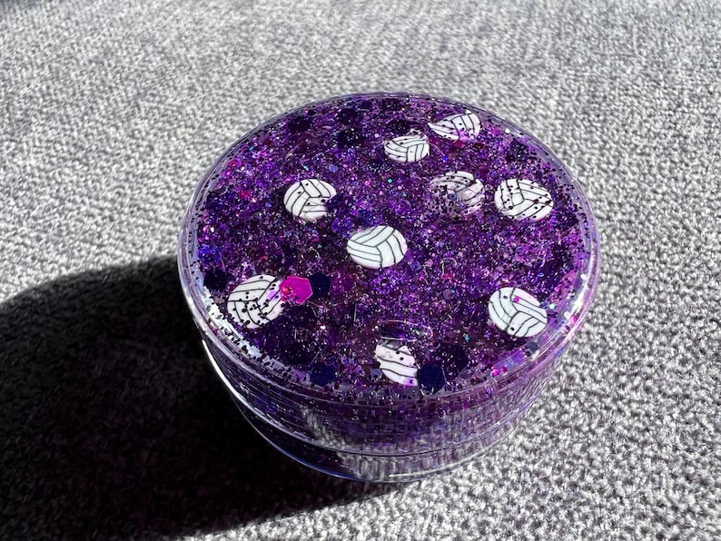 May include: A clear round container filled with purple glitter and white volleyball designs.