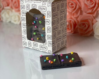 May include: A white box with a black ribbon and a clear window showing a chocolate bar with colorful sprinkles. The box has a repeating pattern of speech bubbles with the words "Hi" and "Follow" in black text. The chocolate bar is sitting on a white surface.