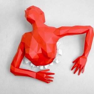 以下が含まれることがあります： 白い表面を突き破る人の赤い紙工作の彫刻。彫刻は多くの小さな三角形でできており、横たわったポーズをとっている。