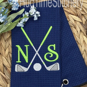 May include: A navy blue golf towel with a black border and a white golf ball, two crossed golf clubs, and the letters "N" and "S" embroidered in green. The towel has a black loop for hanging.
