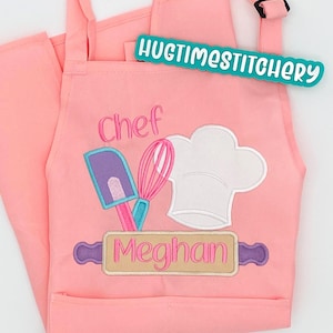 May include: A light pink apron featuring an embroidered design of a white chef hat, a purple and pink spatula, and a pink whisk. Below the tools is a tan rolling pin with the name Meghan embroidered in pink. The text HUGTIMESTITCHERY is visible.