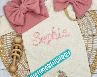 May include: A beige baby onesie with the name "Sophia" embroidered in pink rhinestones. The onesie has a turquoise label that says "Hugtimestitchery". The onesie is paired with a pink bow headband and a pink bow.