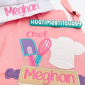 May include: A pink apron and white chef hat set. The apron features embroidered kitchen tools including a purple spatula, pink whisk, and white chef hat, with the name Meghan in pink. The hat also displays the name Meghan in pink embroidery.