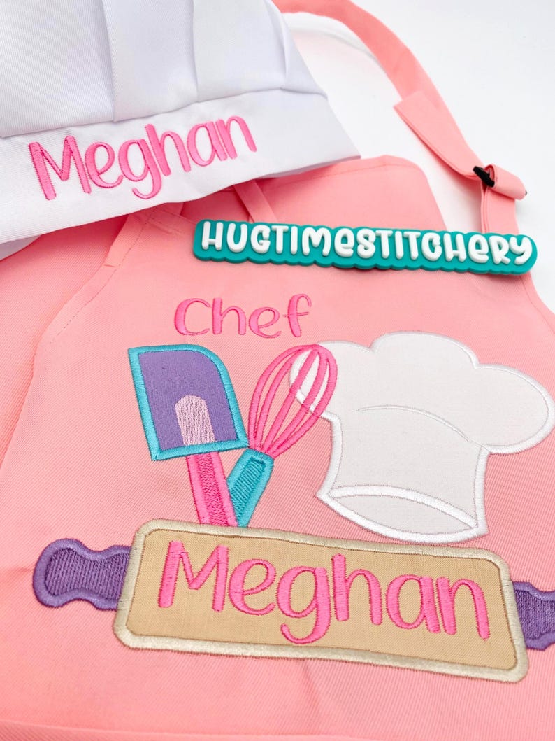 May include: A pink apron and white chef hat set. The apron features embroidered kitchen tools including a purple spatula, pink whisk, and white chef hat, with the name Meghan in pink. The hat also displays the name Meghan in pink embroidery.