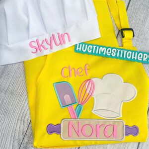 May include: A bright yellow apron and a white chef hat. The hat features the embroidered name Skylin in pink. The apron displays the word Chef in pink, an embroidered graphic of a spatula, whisk, and chef hat, and a rolling pin with the name Nora.