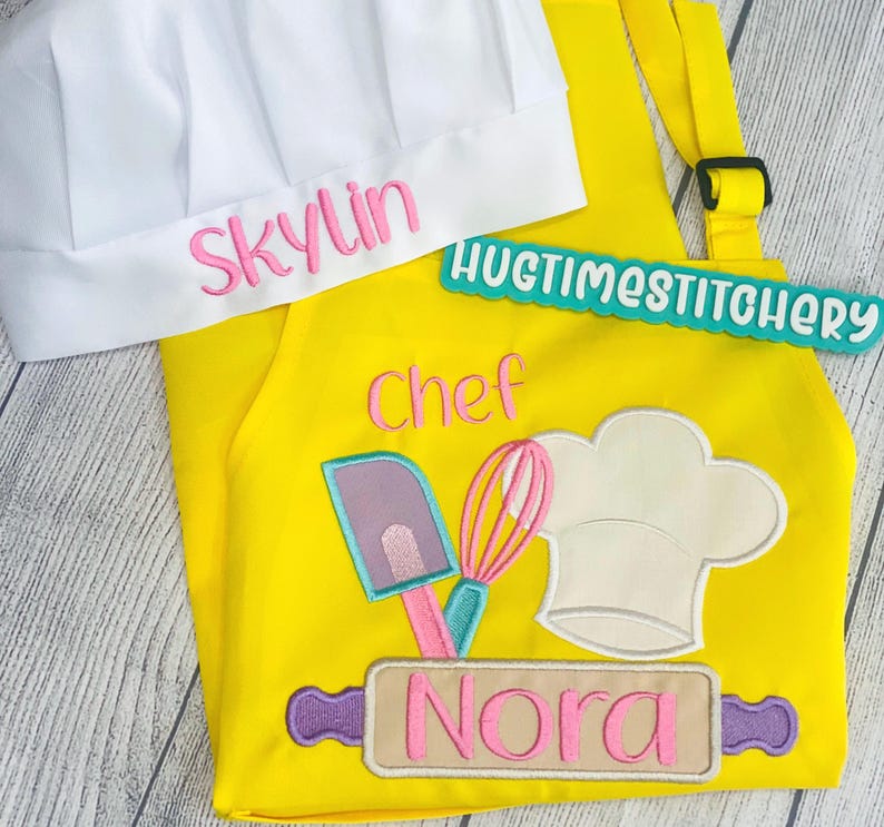 May include: A bright yellow apron and a white chef hat. The hat features the embroidered name Skylin in pink. The apron displays the word Chef in pink, an embroidered graphic of a spatula, whisk, and chef hat, and a rolling pin with the name Nora.