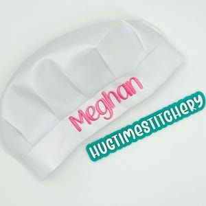 May include: A white chef hat featuring the name Meghan embroidered in pink thread on the front band. Beside the hat is a teal rectangular label with white text that reads HUGTIMESTITCHERY in a rounded, playful font style.