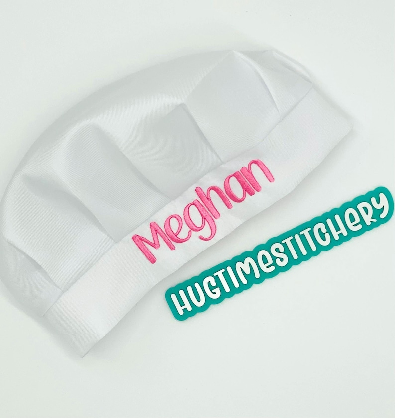 May include: A white chef hat featuring the name Meghan embroidered in pink thread on the front band. Beside the hat is a teal rectangular label with white text that reads HUGTIMESTITCHERY in a rounded, playful font style.