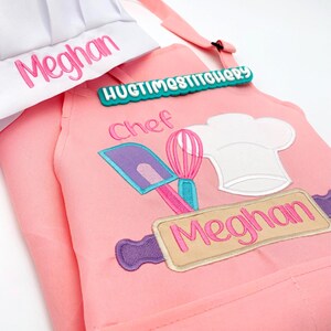 May include: A pink apron and white chef hat set. The apron features embroidered kitchen tools including a purple spatula, pink whisk, and white chef hat, with the name Meghan and the text Chef. A teal tag reads HUGTIMESTITCHERY.