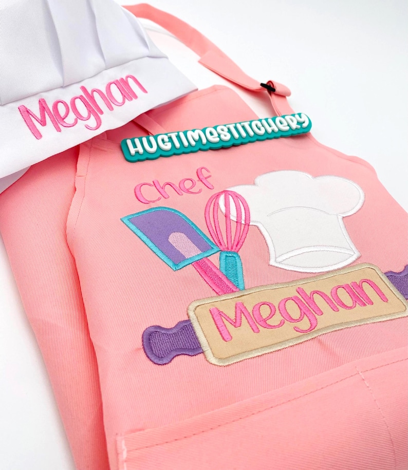 May include: A pink apron and white chef hat set. The apron features embroidered kitchen tools including a purple spatula, pink whisk, and white chef hat, with the name Meghan and the text Chef. A teal tag reads HUGTIMESTITCHERY.