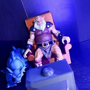 May include: A blue and gray fantasy creature figurine sits in front of a television with a large, gray, bearded, ogre-like figure sitting in a brown chair. The ogre is wearing a brown shirt, a brown belt, and brown pants.