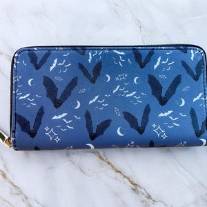 May include: A blue zip-around wallet with a repeating pattern of bats, crescent moons, and stars. The wallet has a gold-colored zipper pull and a black zipper. The design is a dark blue with white accents.