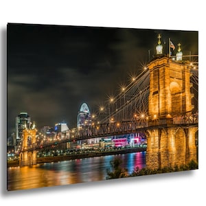 May include: A nighttime cityscape print featuring a suspension bridge illuminated with warm lights. The bridge spans a river, with city buildings and a cloudy sky in the background. The image has a dramatic, artistic feel.