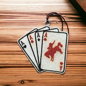 May include: Four white playing cards with glitter, each showing the Ace of a different suit. The cards are arranged in a fan shape, with the Ace of Hearts on top. The Ace of Hearts card features a red glitter silhouette of a cowboy riding a horse.
