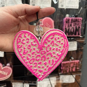 May include: Pink heart-shaped car air freshener with a white leopard print design. The air freshener is hanging from a string with a clear crystal bead.