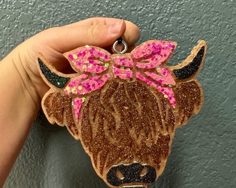 May include: A brown and black Highland cow ornament with a pink glitter bow. The ornament is made of wood and has a glitter finish.