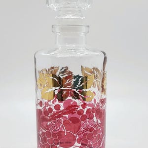 May include: A clear glass decanter with a gold and pink grape design. The decanter has a glass stopper with a ribbed design.