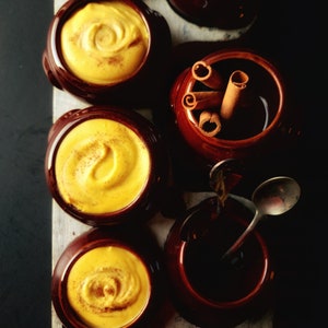 May include: Six small brown ceramic bowls, five filled with a yellow custard-like substance and one with cinnamon sticks. The bowls are arranged on a white wooden surface.