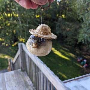 May include: A small, round, white, and brown pufferfish ornament with googly eyes and a straw hat. The ornament is hanging from a string.