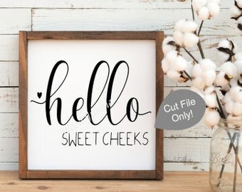 May include: A wooden framed sign with the words "hello sweet cheeks" in black script. A bouquet of cotton stems in a glass jar sits to the right. The background is a white wooden wall.