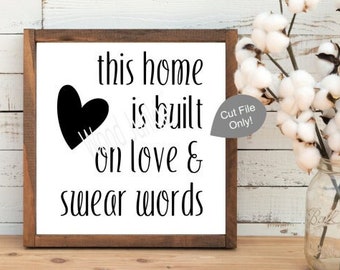 May include: A wooden framed sign with the text "this home is built on love & swear words" in black. A black heart graphic is on the left. A vase of cotton stems is to the right. The sign is on a wooden surface.