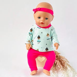 Könnte beinhalten: Eine Puppe trägt ein hellblaues Langarmshirt mit Ballerina-Print, kombiniert mit leuchtend pinkfarbenen Leggings und einem passenden Stirnband. Die Puppe steht auf einer weißen Oberfläche.