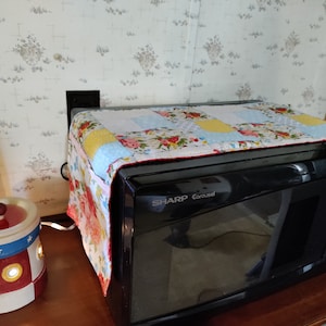May include: A black Sharp Carousel microwave with a colorful patchwork cover. The cover features floral and geometric patterns in shades of red, yellow, blue, and white. A decorative lighthouse-shaped lamp sits to the left.