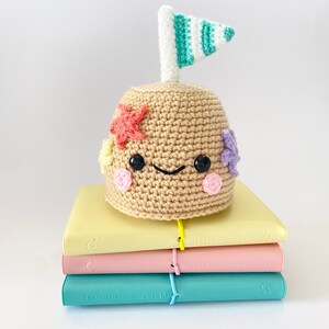 May include: A crocheted sandcastle toy with a smiling face, decorated with colorful starfish and flowers. The sandcastle has a small flag on top and is sitting on top of a stack of colorful notebooks.