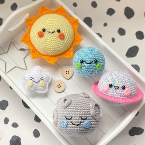 May include: A selection of crocheted celestial bodies, including a smiling sun, planet Earth, Saturn, a star, and a moon. The sun is yellow with orange rays, and the planet is blue and green. The star is white, and the moon is grey.