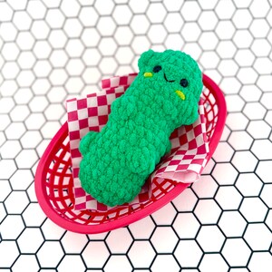 May include: A green crocheted plush toy shaped like a pickle, with a smiling face, lying in a red and white checkered paper-lined red plastic basket.