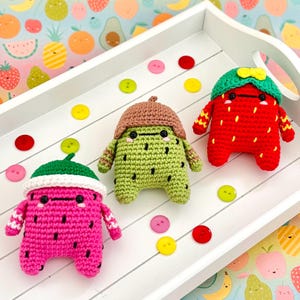 Pode incluir: Três brinquedos de pelúcia de crochê em forma de melancia, morango e abacate. Cada brinquedo tem um rosto bonito e está usando um chapéu. Os brinquedos estão sentados em uma bandeja branca com um fundo colorido.