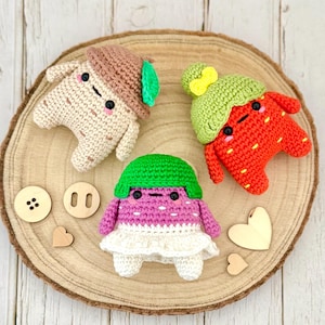 May include: Three crocheted stuffed toys in the shape of a mushroom, strawberry, and a purple character with a green hat. The toys are sitting on a wooden surface.