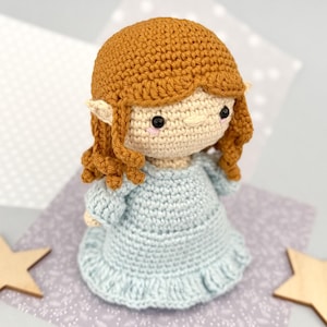 May include: A handmade crochet doll with a light blue dress and auburn hair. The doll has a cream-colored face with black eyes and pink cheeks. The doll's hair is styled in a curly bob. The doll is standing on a patterned surface.