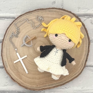 May include: A handmade crochet doll with a yellow head of hair, a white dress, and a black jacket. A silver cross necklace and a miniature crossbow are also on display, all arranged on a wooden slice.