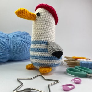 May include: A crocheted seagull toy with a red and white striped hat, blue and white striped shirt, and grey and black wings. The toy is sitting on a white surface.