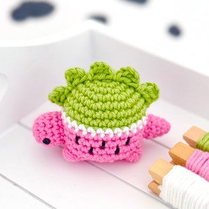 May include: A pink and green crocheted dinosaur toy with a white trim. The toy is sitting on a white surface with wooden spools of yarn.